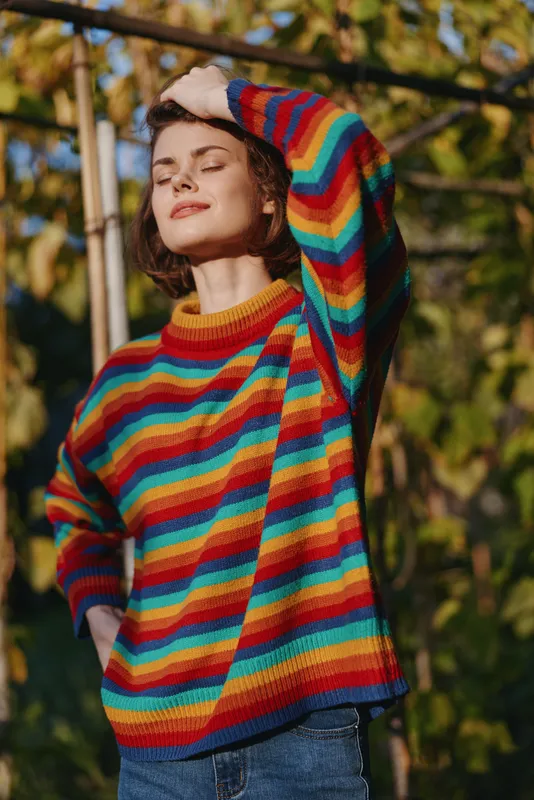 Woman striped sweater casual outfit stands outdoors in sunlight garden, smiling with eyes closed and hand on head, relaxed young adult in denim jeans for lifestyle fashion portrait.