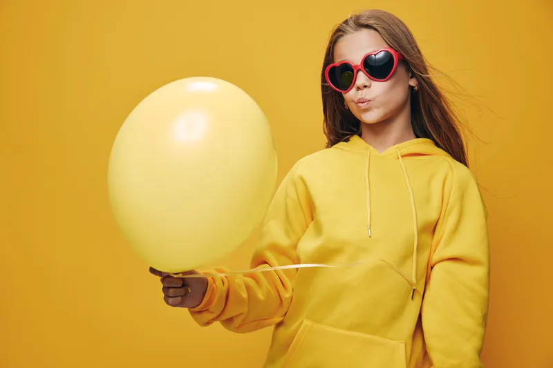 Girl wearing red heart shaped sunglasses and yellow hoodie holding a yellow balloon on bright yellow background. Fun, casual style with playful expression and vibrant colors.