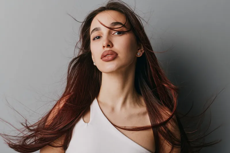 Young woman with long flowing hair and natural makeup posing in white top against gray background. Beauty portrait with confident expression and wind effect on hair in studio lighting.