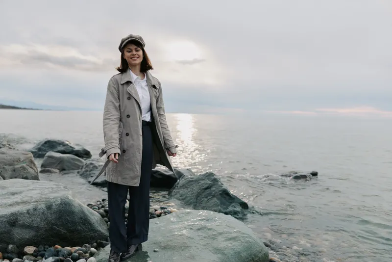 Smiling woman in casual coat and pants stands on rocky beach near calm sea under cloudy sky. Concept of outdoor lifestyle, autumn fashion, and peaceful nature scene.