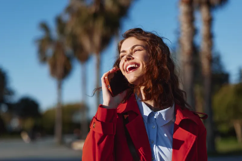Happy woman with curly hair talking on smartphone outdoors in sunny park. She wears red coat and blue shirt, enjoying conversation and bright daylight. Casual lifestyle.