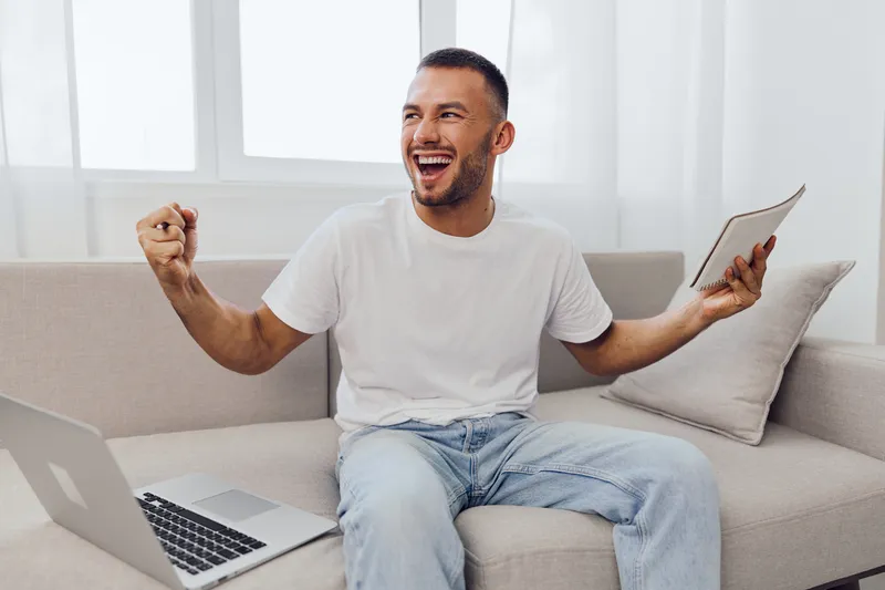 Happy man celebrating success at home with a laptop and notebook, showcasing enthusiasm and productivity in a cozy environment. Enjoying a vibrant and modern lifestyle.