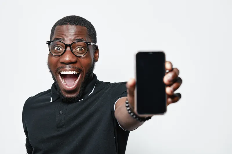 African American man with dark skin and glasses showing smartphone with blank screen excitedly in studio. Black male wearing black polo shirt smiling with open mouth holding phone closeup.