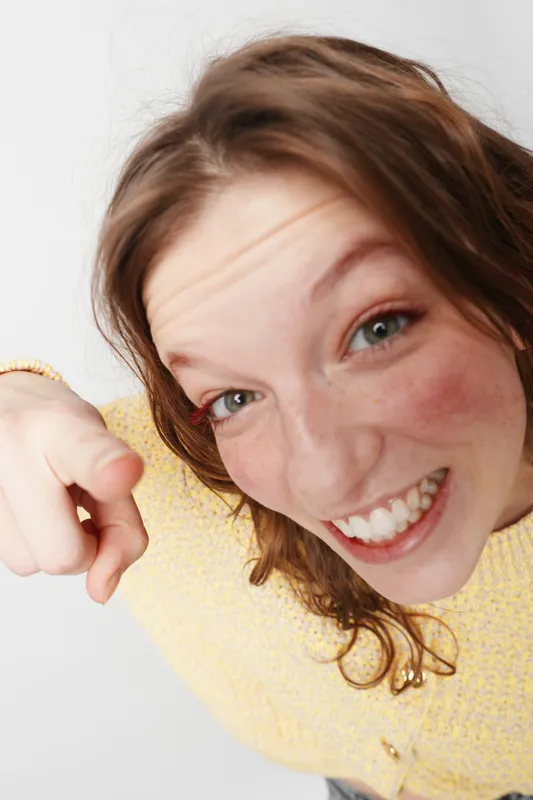 Happy young woman with curly hair wearing yellow sweater smiles brightly and points at camera. Close-up portrait on white background. Concept of joy, friendliness, and positive energy.
