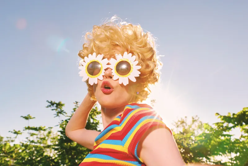 A woman in a striped rainbow top poses outdoors with oversized daisy sunglasses, capturing a playful retro lifestyle vibe and sunny nostalgia.