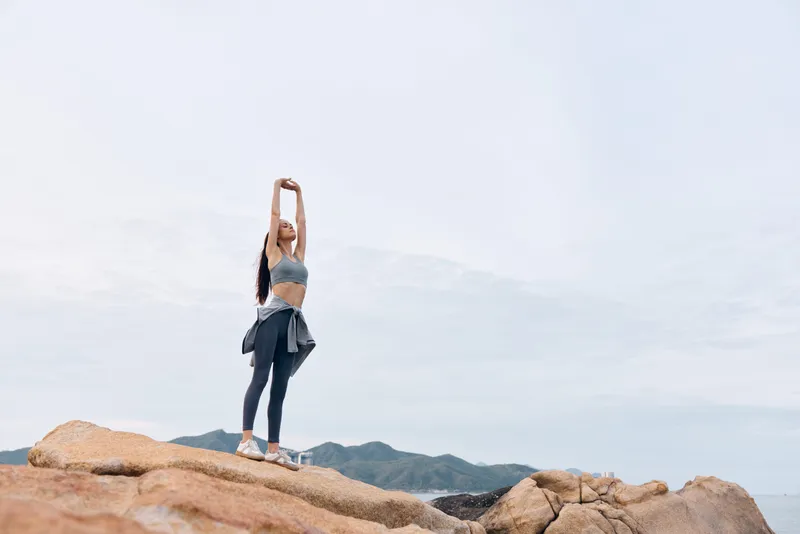 Young woman stretching outdoors on rocks, wearing athletic sportswear and enjoying a moment of tranquility in nature, embodying health and fitness concepts