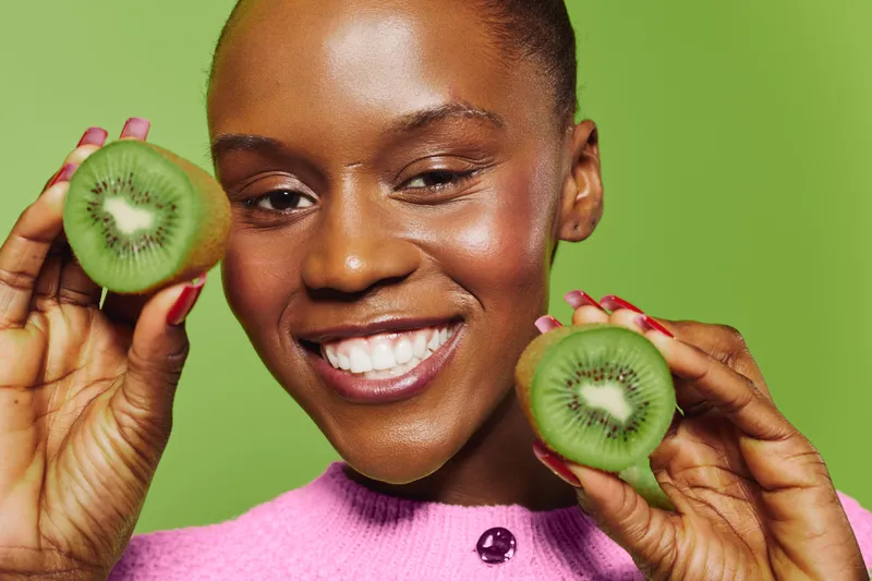Happy African woman with dark skin holds two halves of fresh kiwi fruit smiling against green background. Concept of healthy lifestyle, natural food, and positive emotions in wellness.