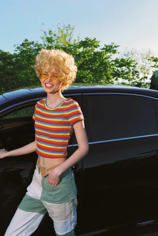 Woman in a striped crop top and patchwork jeans stands beside a sleek car, enjoying a sunny day, retro film color mood, candid lifestyle portrait outdoors