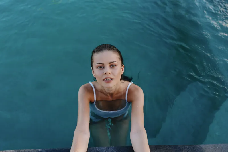 Young woman in blue swimsuit climbing out of turquoise water, looking up with wet hair, natural light, summer lifestyle, outdoor swimming pool scene, relaxed expression