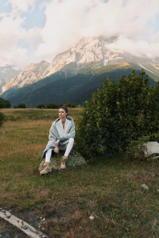 Young woman lifestyle traveler sits on grass near bush in mountain valley wrapped in blanket enjoys peaceful nature landscape with cloudy sky and rocky peaks in background