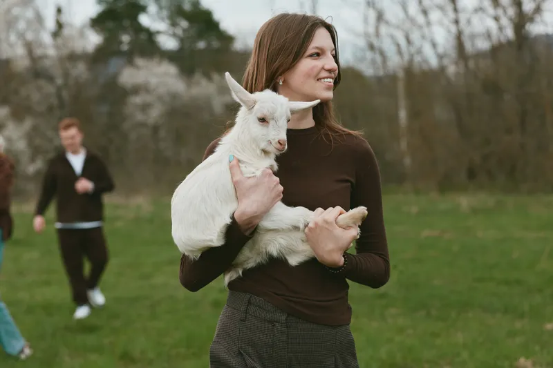 Young woman holds a small white baby goat in her arms outdoors during a casual lifestyle moment in early spring with a soft smile and relaxed posture