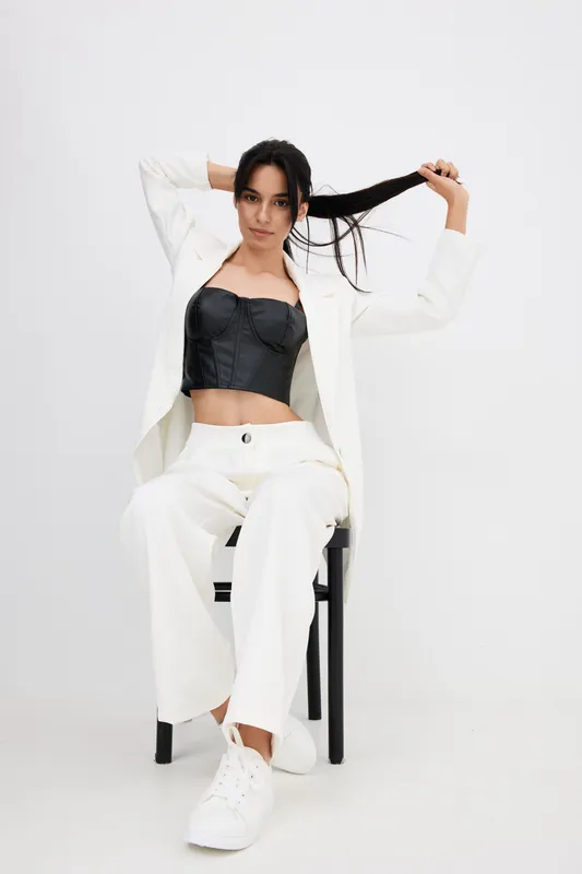 Young woman in white suit and black corset sitting on chair, holding her dark hair playfully, minimalist studio background with casual confident pose.