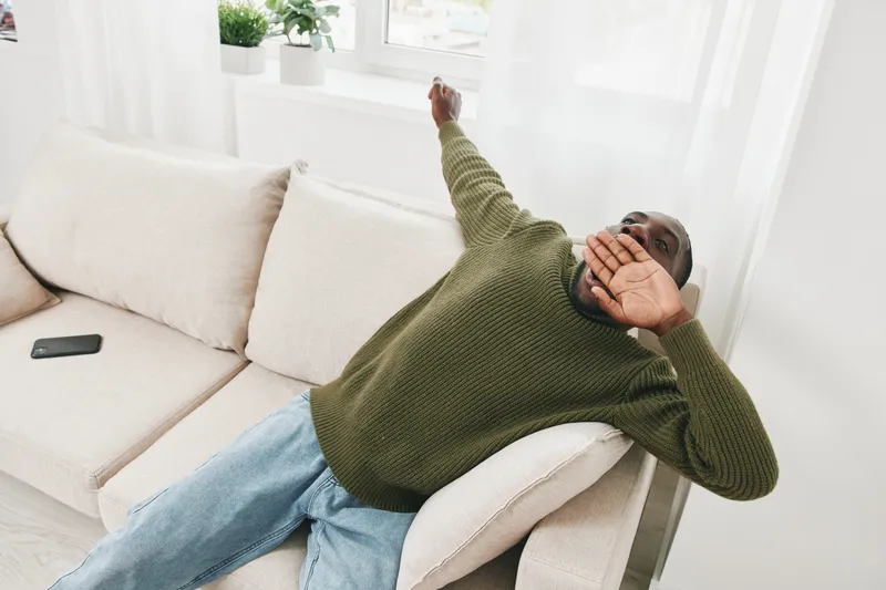 A Black man relaxing on a light sofa at home, wearing a cozy green sweater and blue jeans, expressing comfort and tranquility in a bright, airy space