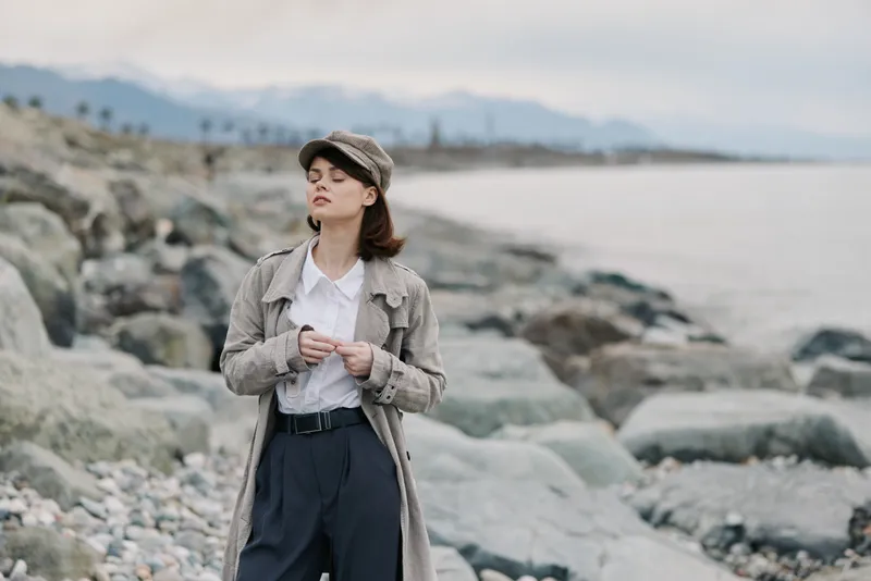 Young woman in vintage outfit stands on rocky shore near calm sea under cloudy sky. She wears textured coat and cap, eyes closed, enjoying peaceful coastal atmosphere and fresh air.