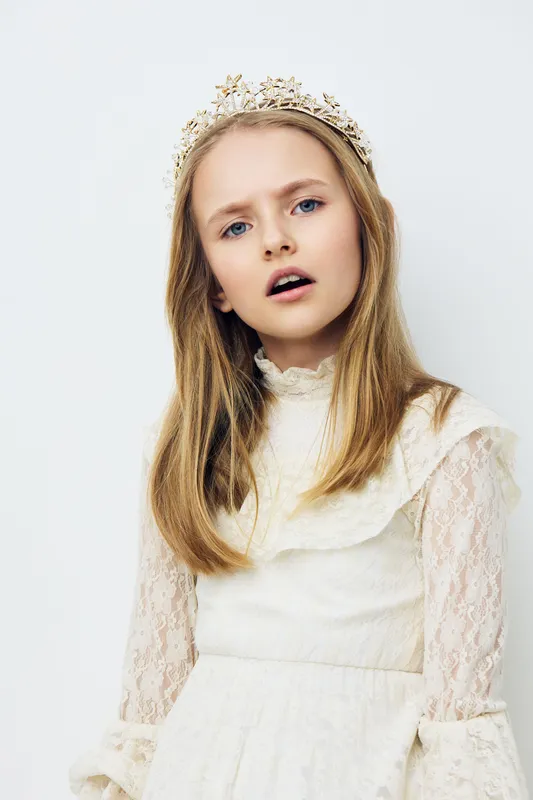 Portrait of a young girl with long blonde hair wearing a delicate crown, dressed in a light dress, conveying innocence and purity in a soft, minimalist setting