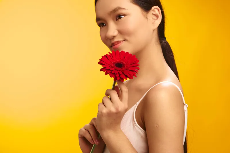 Asian woman smiles softly as she holds a red flower in a bright studio, creating a vivid, warm portrait with a calm and inviting mood, perfect for lifestyle and beauty projects.