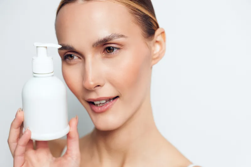 Young woman with clear skin holds white cosmetic bottle in hand looks at product with gentle smile isolated on white background for skincare concept and beauty routine.