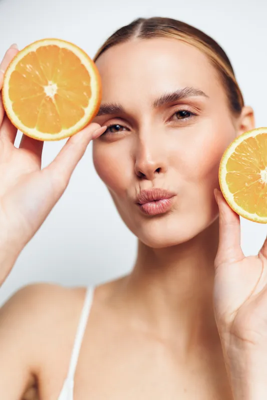 Young woman holding two orange halves near face, making a playful kiss expression, fresh citrus fruit, natural skincare concept, bright background, close-up portrait