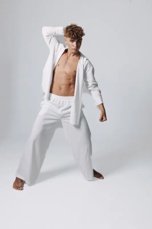 Young man posing topless in white pants and shirt with curly hair on white background. Expression, casual look, dynamic pose with contemporary fashion style and confident attitude.
