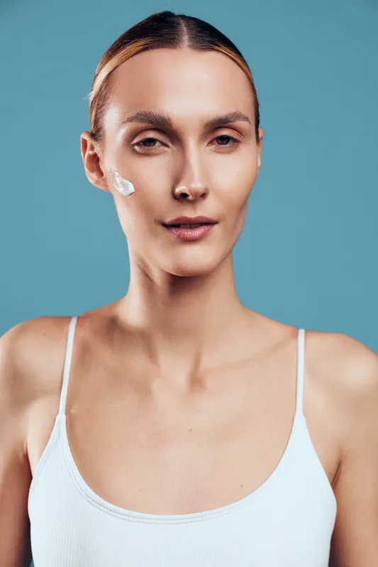 Young woman with clear skin and cream on cheek, wearing white tank top, looking confidently at camera, smooth complexion, blue background, minimal makeup, skincare concept