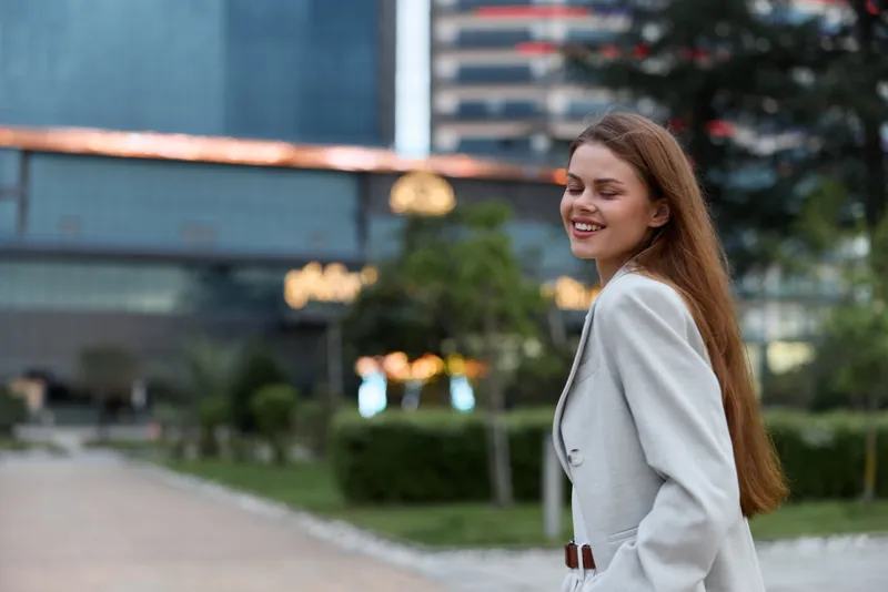 Smiling woman in light gray business suit walking outdoors in urban environment with modern buildings and greenery, expressing confidence and professionalism