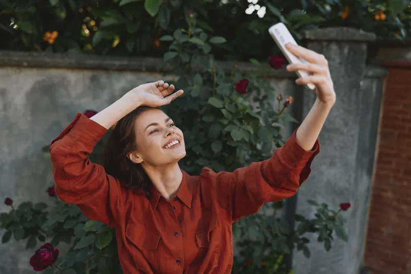 Cheerful woman outdoors, taking a selfie with a bright smile, red shirt, casual pose, natural light, soft breeze, lush greenery background, candid lifestyle moment, relaxed vibe