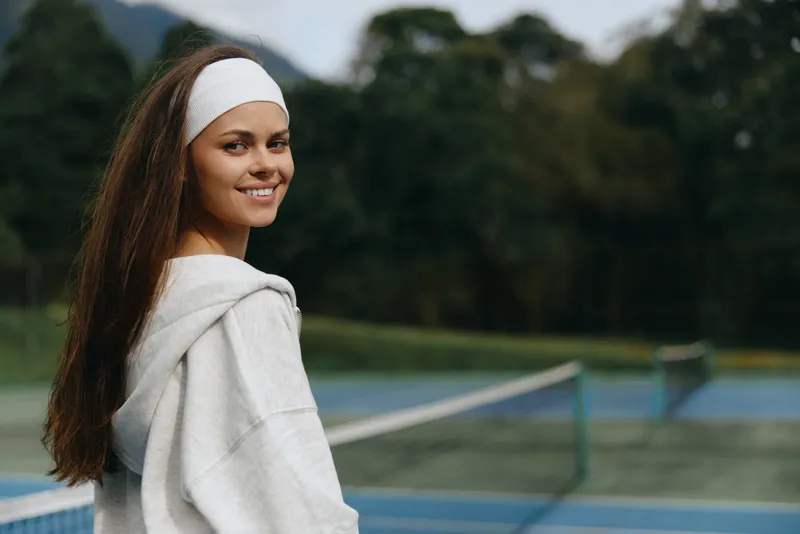 Young female tennis player smiling on the court with a relaxed outfit, showcasing happiness and confidence against a lush green background