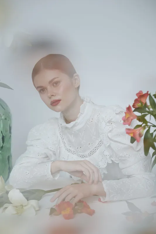 Elegant young woman in white dress with lace details, featuring a serene expression, surrounded by flowers and a soft pastel background in a spring aesthetic
