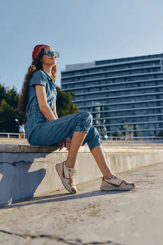 Lifestyle woman in modern boho-western denim street style sitting outdoors on sunny day, wearing sunglasses and a red bandana, film color vibe