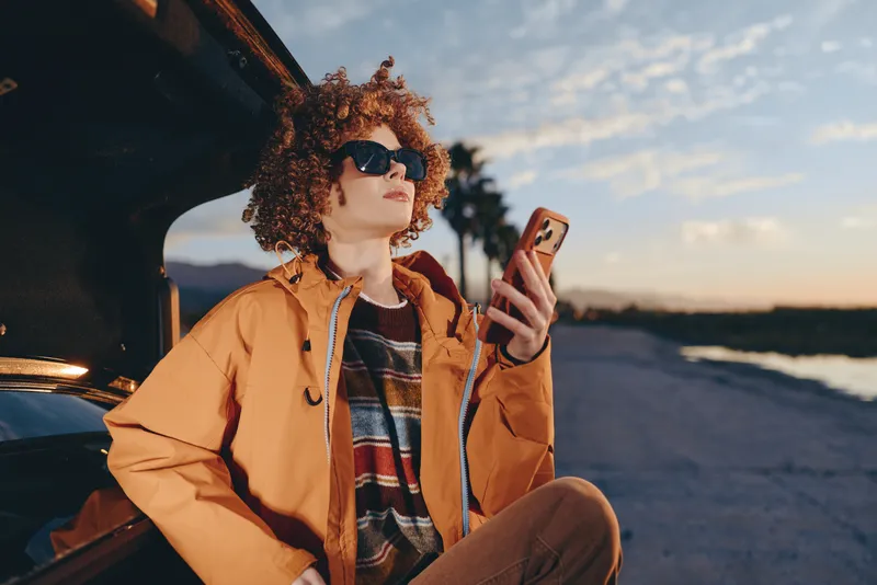 Woman lifestyle curly hair sunglasses smartphone car sunset outdoors portrait, sitting on curb beside car outdoors at sunset, holding smartphone and scrolling, warm golden light, casual travel mood