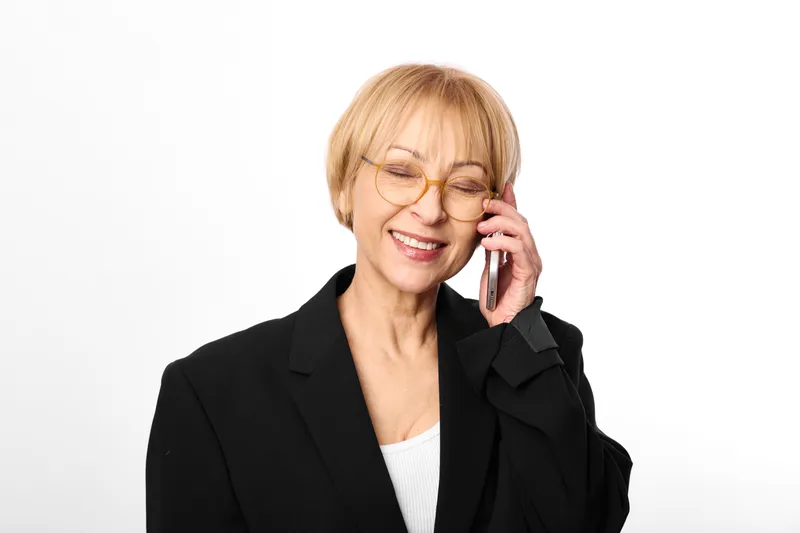Elderly woman in business blazer, smiling while talking on phone, wearing glasses, short blond hair, friendly expression, clean white background, studio portrait