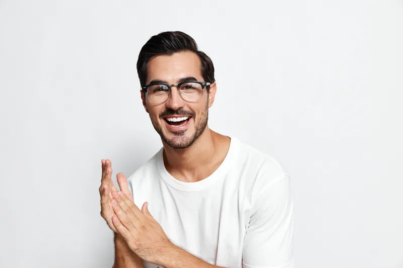 Smiling man with glasses wearing white t-shirt clapping hands in excitement, showcasing joy and enthusiasm against a minimalistic light background.
