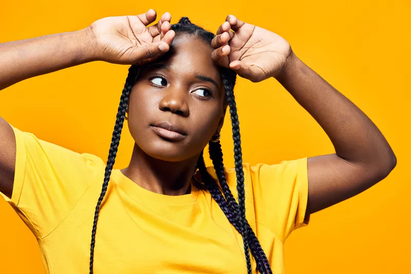 A young African woman with braids poses against a vibrant yellow background, showcasing confidence and strength in a casual yellow tee, perfect for wellness themes