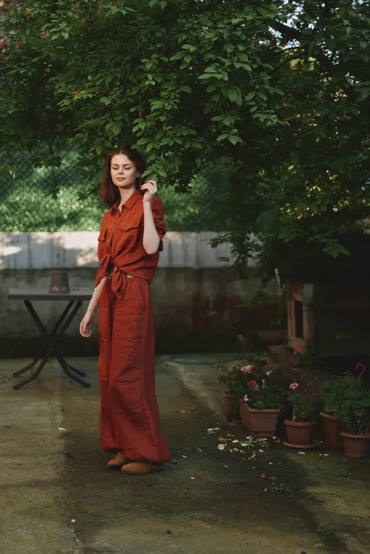 Young woman in a red dress walks through a shaded garden, hair loose, a soft breeze lifting the hem, capturing a relaxed lifestyle moment outdoors with lush greenery