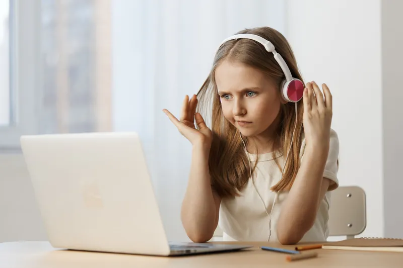 A young girl wearing headphones, engaged in an online lesson, displaying concentration and determination in a bright, modern workspace with a laptop
