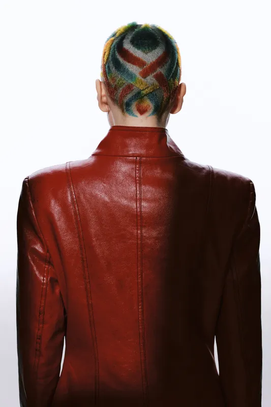 Woman with colorful shaved hair design, wearing a shiny red leather jacket, photographed from behind against a white background, showcasing bold fashion and creativity.