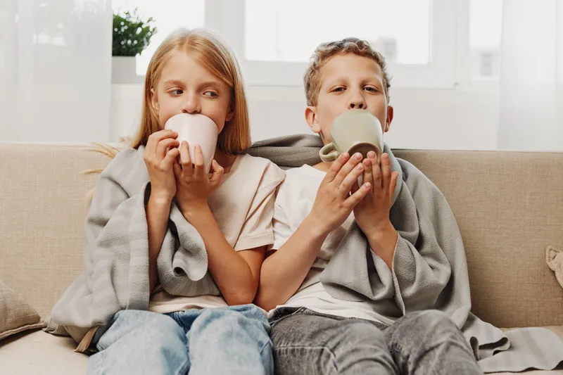 Two children wrapped in cozy blankets sit together on a couch drinking warm beverages from mugs in a bright home setting, enjoying comfort and calm moments.