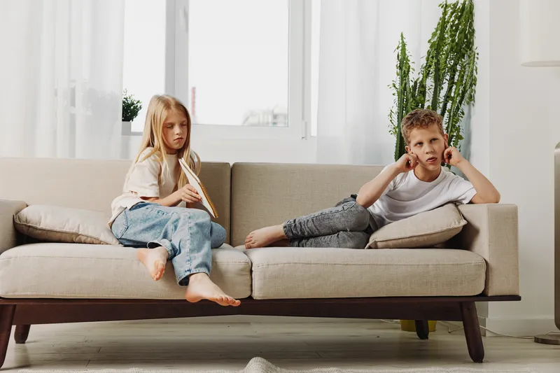 Two bored children sit on a beige sofa indoors, a girl holding a book and a boy covering his ears. The bright living room features plants and natural light from large windows.
