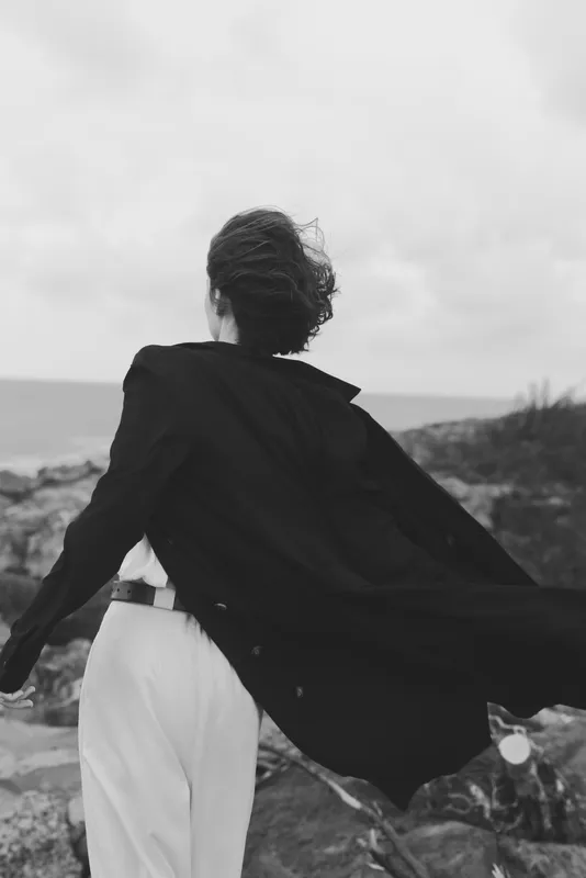 Cape fluttering, flowing cloak behind a figure moving toward the windy coast, white trousers, dark top, monochrome scene, ocean on the horizon, dramatic movement and resilience, minimal styling,