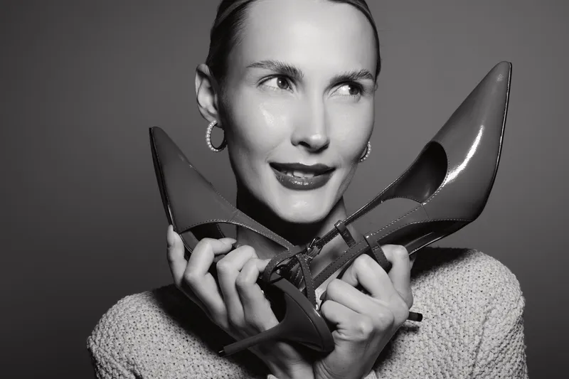 Portrait of a smiling Caucasian woman with light skin holding two high heel shoes near her face. Studio shot in black and white with soft lighting. Concept of fashion and elegance.