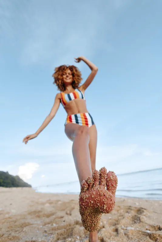 Woman dancing on the beach in summer, curly hair moving in the breeze, joyful smile, striped swimsuit, sunlit ocean shoreline, blue sky with clouds, relaxed pose, outdoor vacation lifestyle