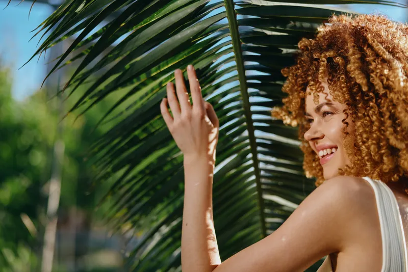 smiling woman with curly hair enjoys tropical sunlight near palm leaves, showing natural beauty and warmth in a bright outdoor setting