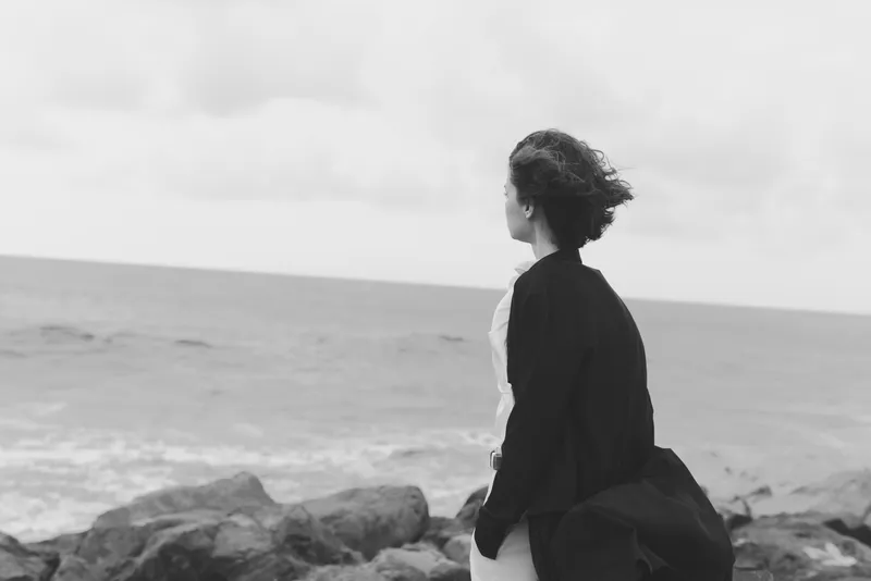 Woman standing on rocky coast, wind in her coat, looking toward the sea, solitary figure captures mood of contemplation, sea breeze, open horizon, monochrome imagery, thoughtful moment by the