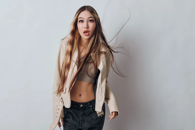 Young woman with long hair and casual outfit posing with wind effect on hair against plain background in studio. Female model wearing crop top and jacket.