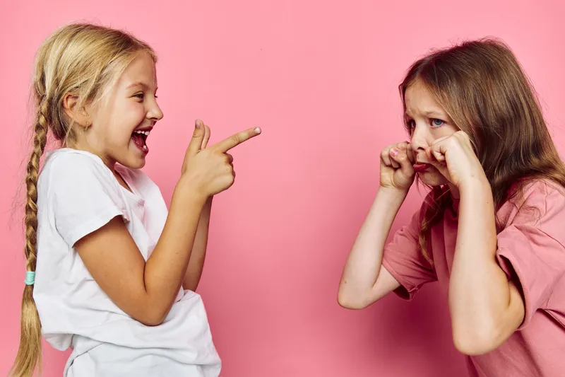 Two young girls interact in front of a pink background one girl laughs and points while the other pretends to cry with fingers by her eyes.