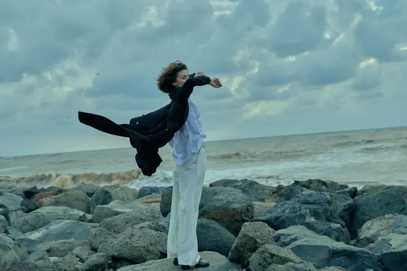 Woman on rocky coast with scarf flowing in wind by the sea, outdoor fashion scene, dramatic movement, serene sky, white dress and dark scarf, natural maritime setting