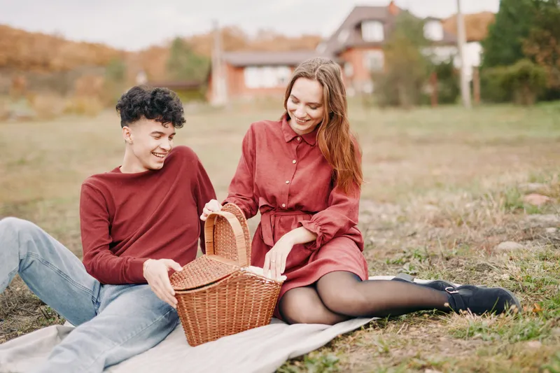 A candid scene captures authenticity as two friends share a relaxed picnic on a sunlit field, with genuine smiles, cozy posture, and a woven basket resting nearby.