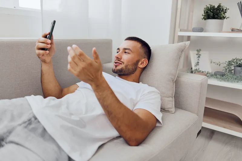 Relaxed man enjoying a video call while lounging on his couch, dressed casually in a white t-shirt. The modern interior creates a cozy atmosphere, reflecting contemporary lifestyle trends.