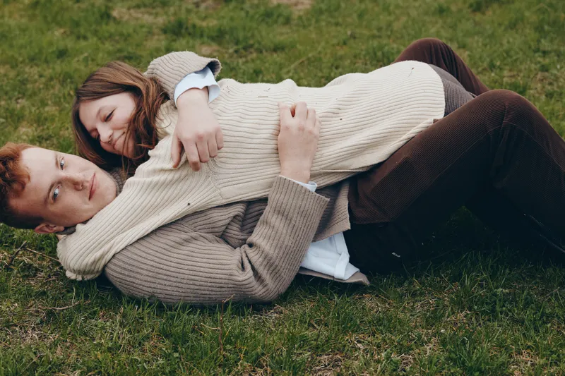 A couple embraces on the grass in cozy sweaters, enjoying an outdoors moment that captures affection, warmth, and connection in a relaxed lifestyle scene.