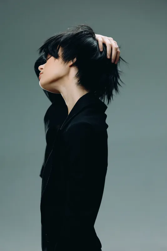Young woman with short black hair styled in a modern cut wearing a black blazer in a minimalistic studio setting.